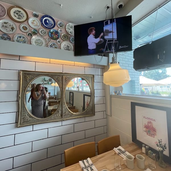 A restaurant interior with decorative plates, a TV showing Bob Ross, mirrors, and a set table.