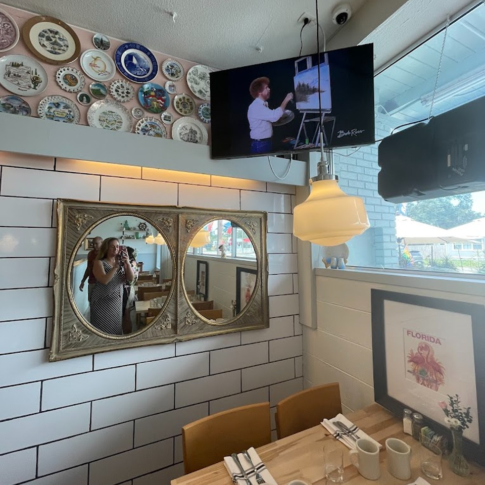 A restaurant interior with decorative plates, a TV showing Bob Ross, mirrors, and a set table.