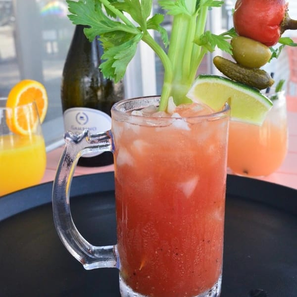 A Bloody Mary cocktail with celery, lime, olive, pickle, and pepper garnishes, next to an orange juice.
