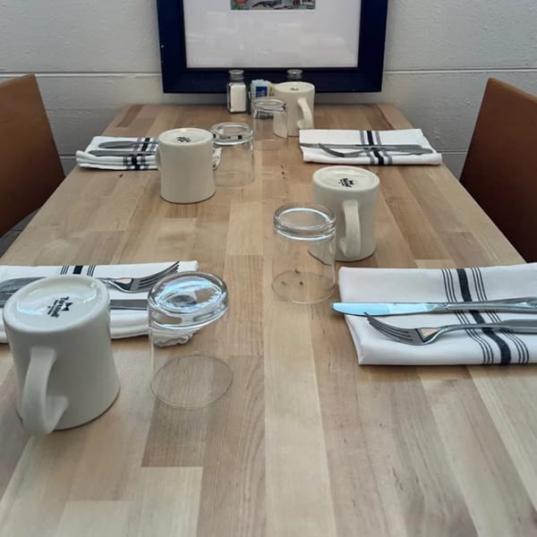 A wooden dining table set for four with white mugs, glasses, and striped napkins with silverware.