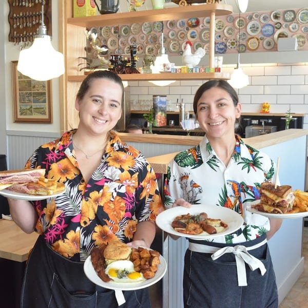 Two smiling servers in Hawaiian shirts hold plates of American breakfast and lunch dishes at Tchotchke.