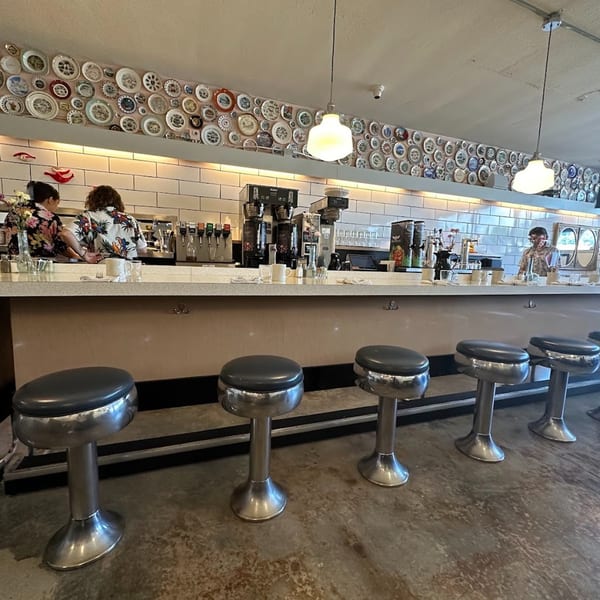 Diner-style counter with chrome stools and two staff members, decorated with many plates on the wall.